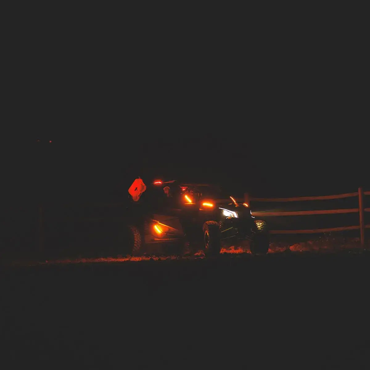 Off-road vehicle with headlights on at night near a wooden fence.