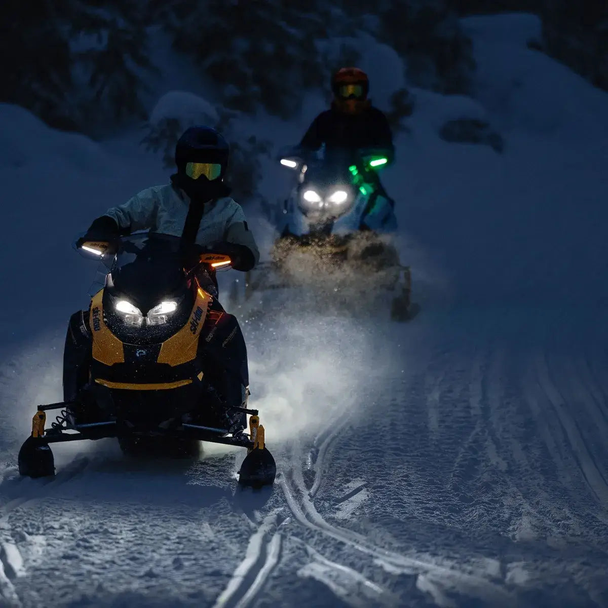 Two snowmobilers on a snowy trail at night with headlights illuminating the path.