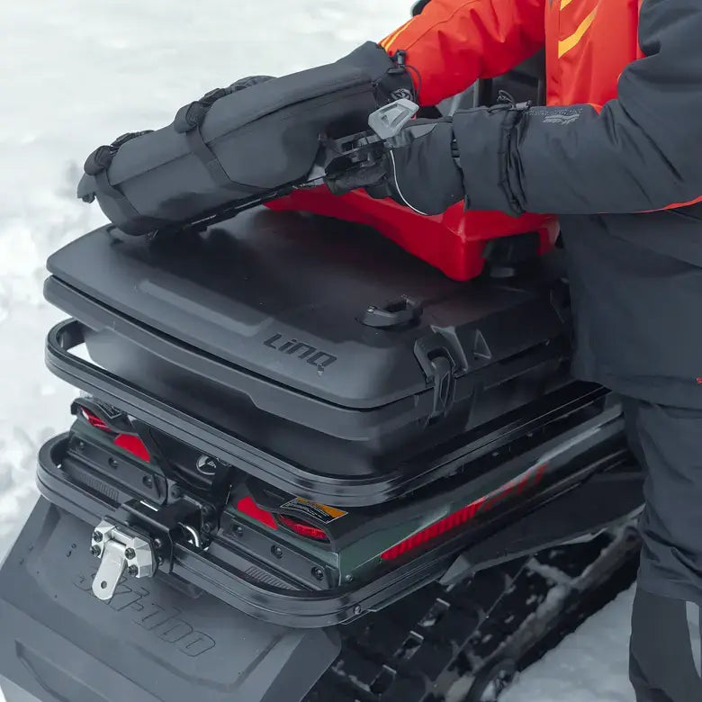 Person adjusting a black snowmobile carrier on a snowmobile in a snowy environment