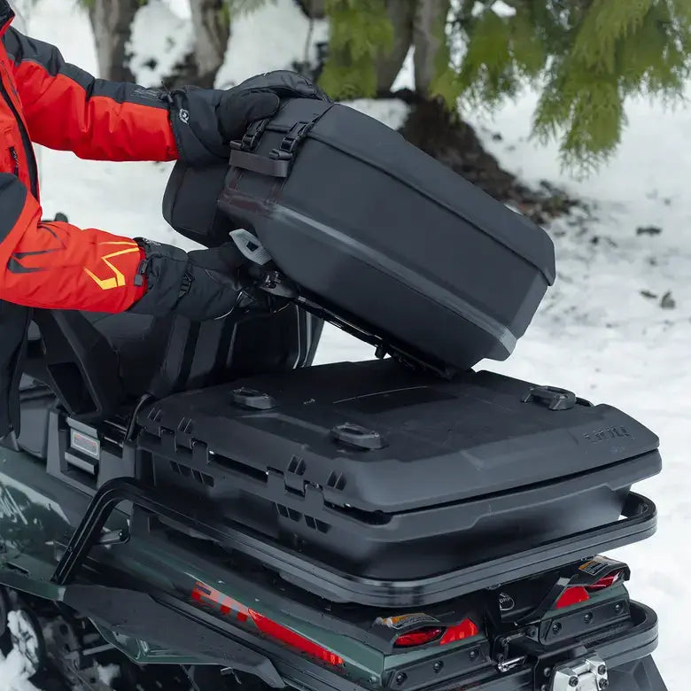 Person in red jacket and black gloves attaching a black storage box to a vehicle in a snowy setting.