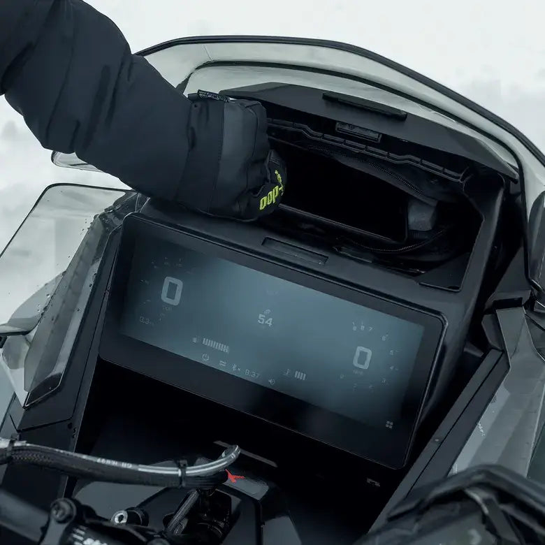 Close-up of a motorcycle's digital display with a gloved hand interacting with it, set against a snowy background.