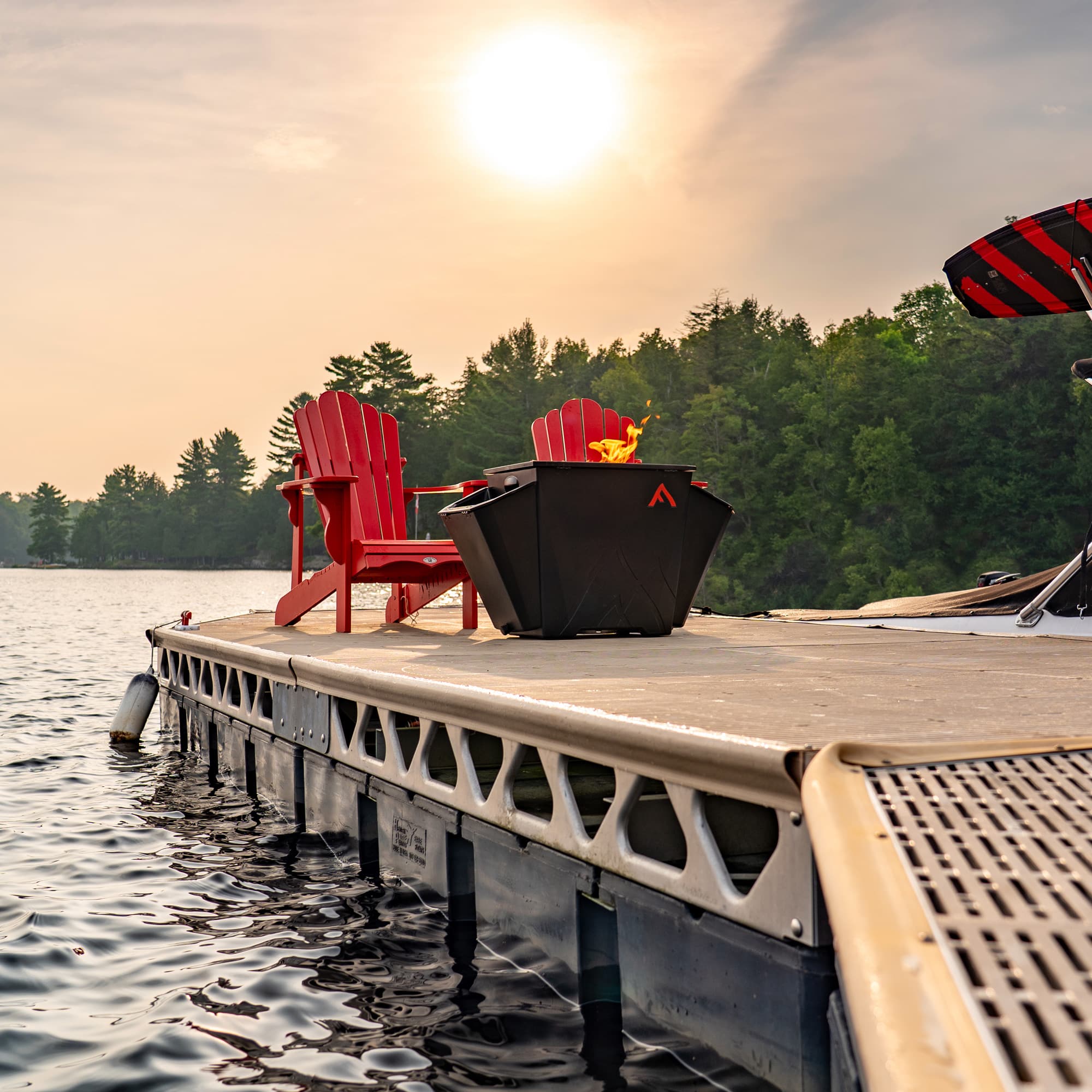 Floating Flame portable fire pit burning on dock beside red Muskoka chairs by lake