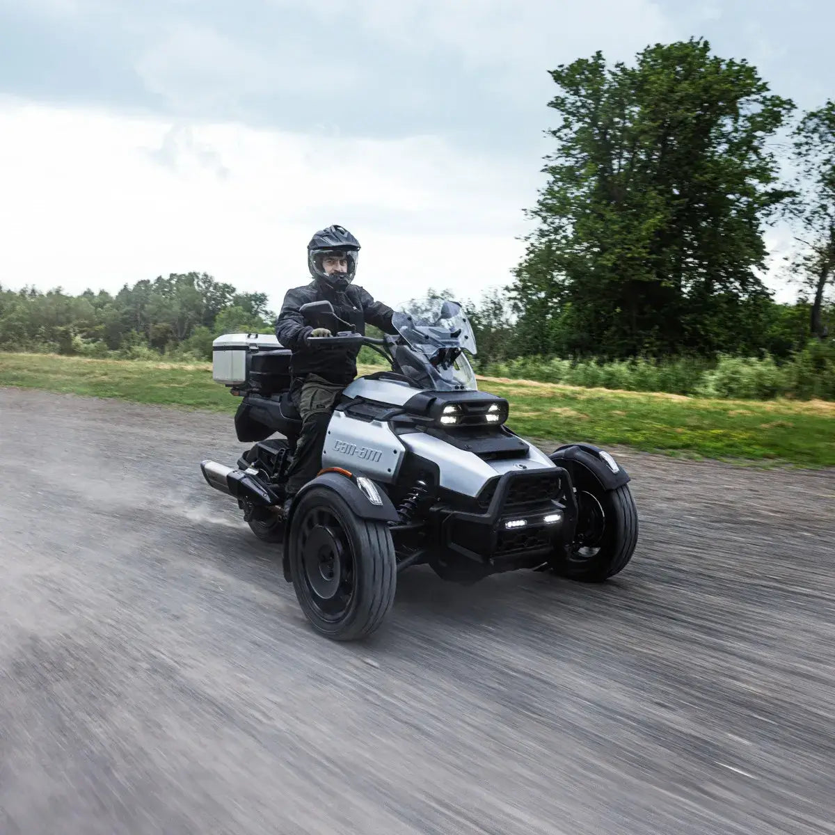 Person riding a BRP Canyon three-wheeled vehicle on a road with trees in the background