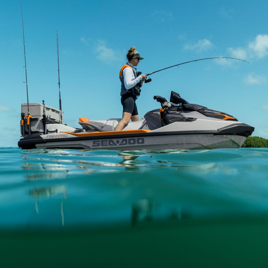 A person stands on a Sea-Doo jet ski fishing in clear blue water with a shoreline and trees visible under a bright sky.