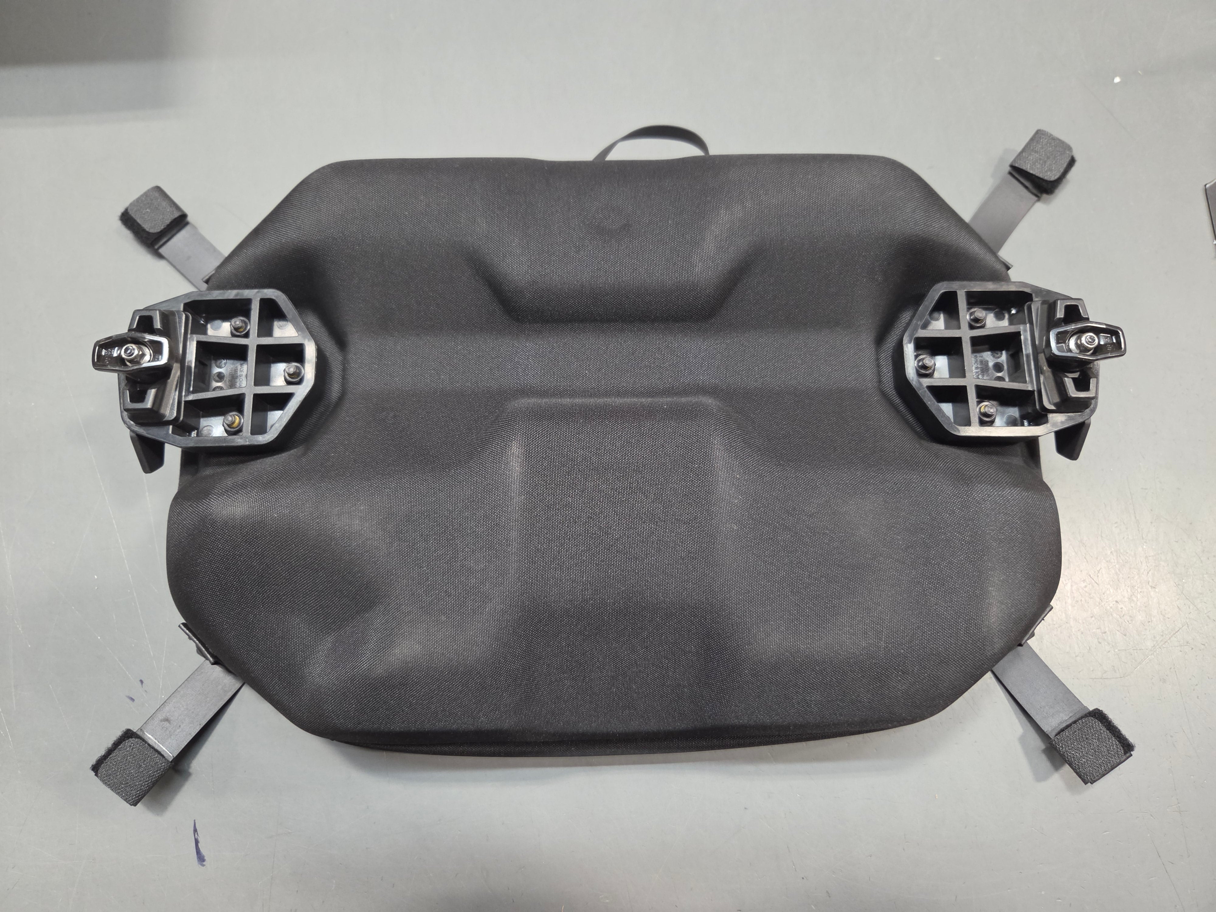 A dark gray molded plastic automotive part, likely a hood ornament or splash shield, with four mounting clips attached to the corners, photographed on a light gray surface.
