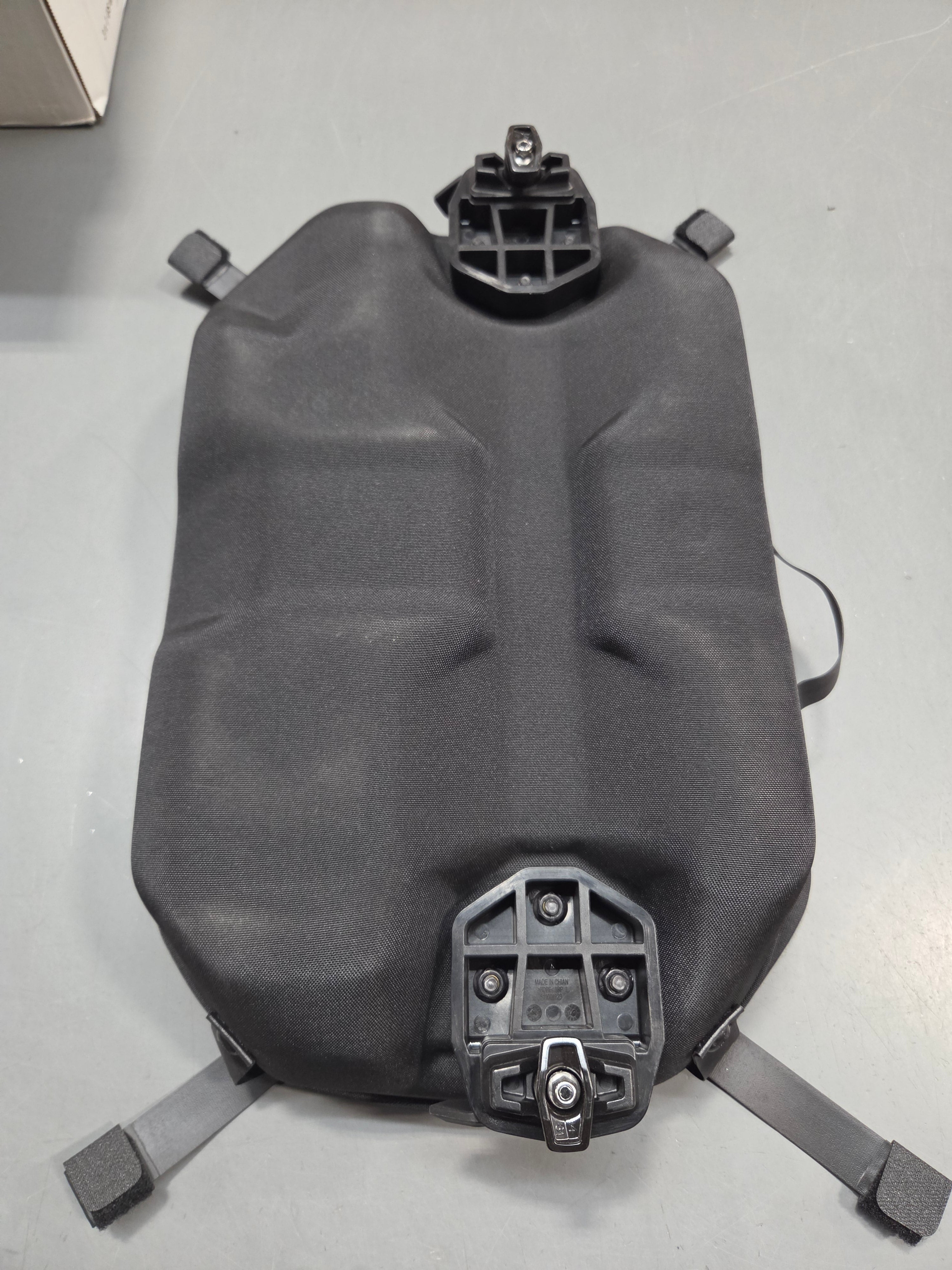 A dark gray molded plastic seat or cushion with integrated mounting hardware and straps, photographed from above on a light gray surface.