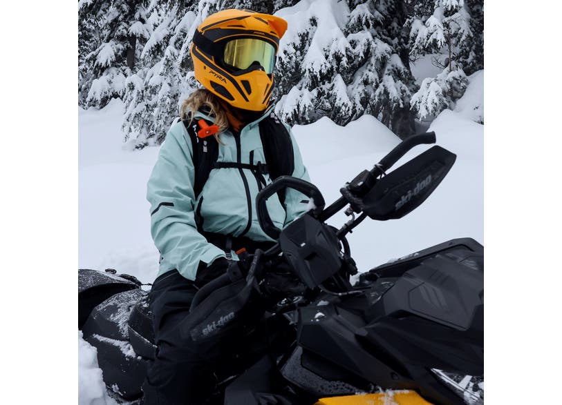 Rider on Ski-Doo snowmobile wearing black flexible handguards mounted on handlebars, snowy forest backdrop