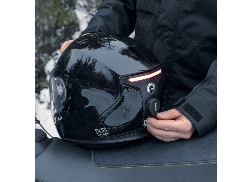 Rider holding glossy black Ski-Doo M-Forge helmet with E-LinQ module, side view on snowmobile seat