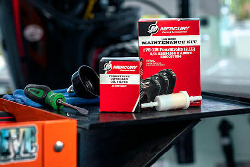 Mercury engine maintenance kit components, including a red and white box and a small white tube, displayed on a black workbench with tools and a blurred engine in the background.