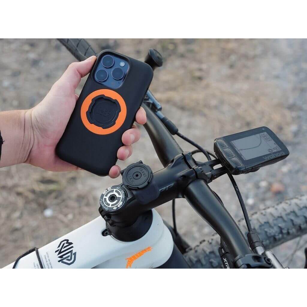 Hand holding a phone with an orange ring mount attached to a bicycle handlebar.